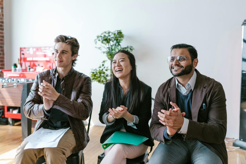 Three diverse team members clapping and smiling during an indoor meeting, showing positivity and teamwork.